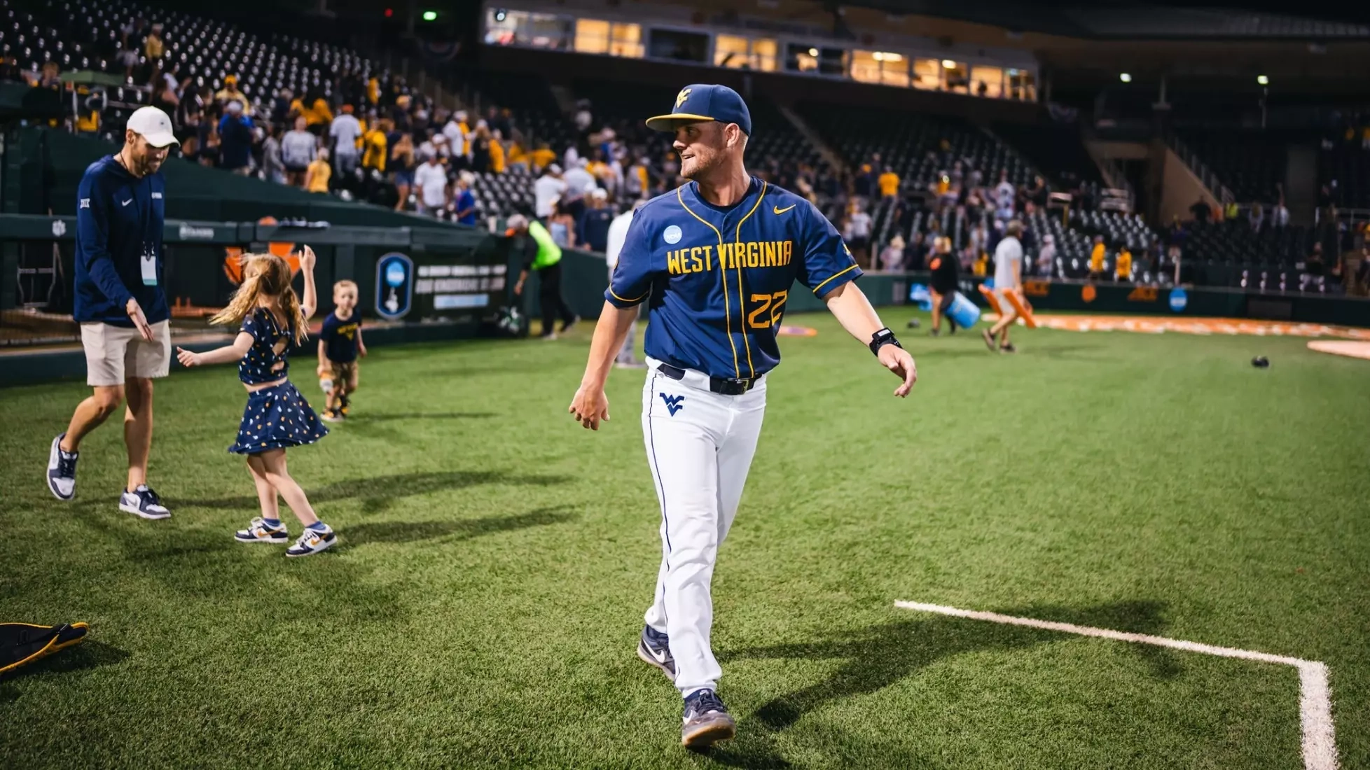 West Virginia baseball player walking on the field after a game, with fans and children playing in the background.