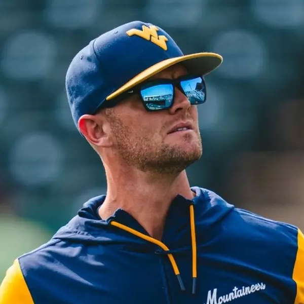 Steve Sabins wearing a West Virginia Mountaineers cap, sunglasses, and blue-and-gold athletic top stands on a baseball field with blurred stadium seating behind him.