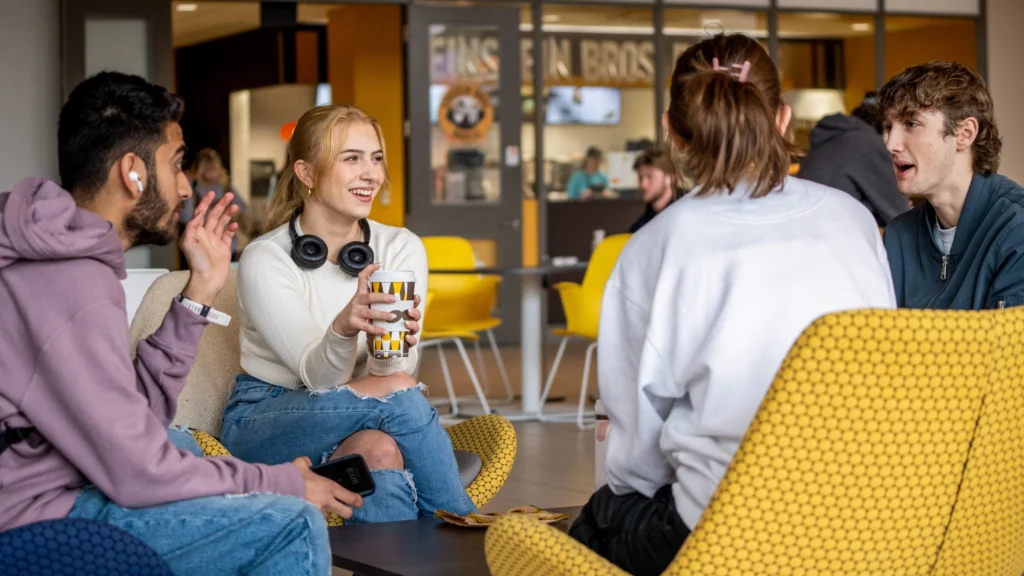 Four college students sit in a lounge area, talking and smiling; one holds a drink while another gestures, with a café and other students in the background.