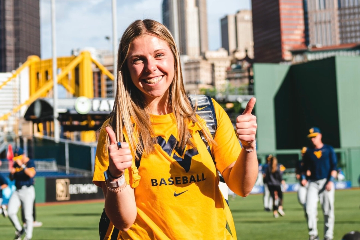 A woman in a West Virginia baseball shirt smiles and gives two thumbs up on a baseball field.