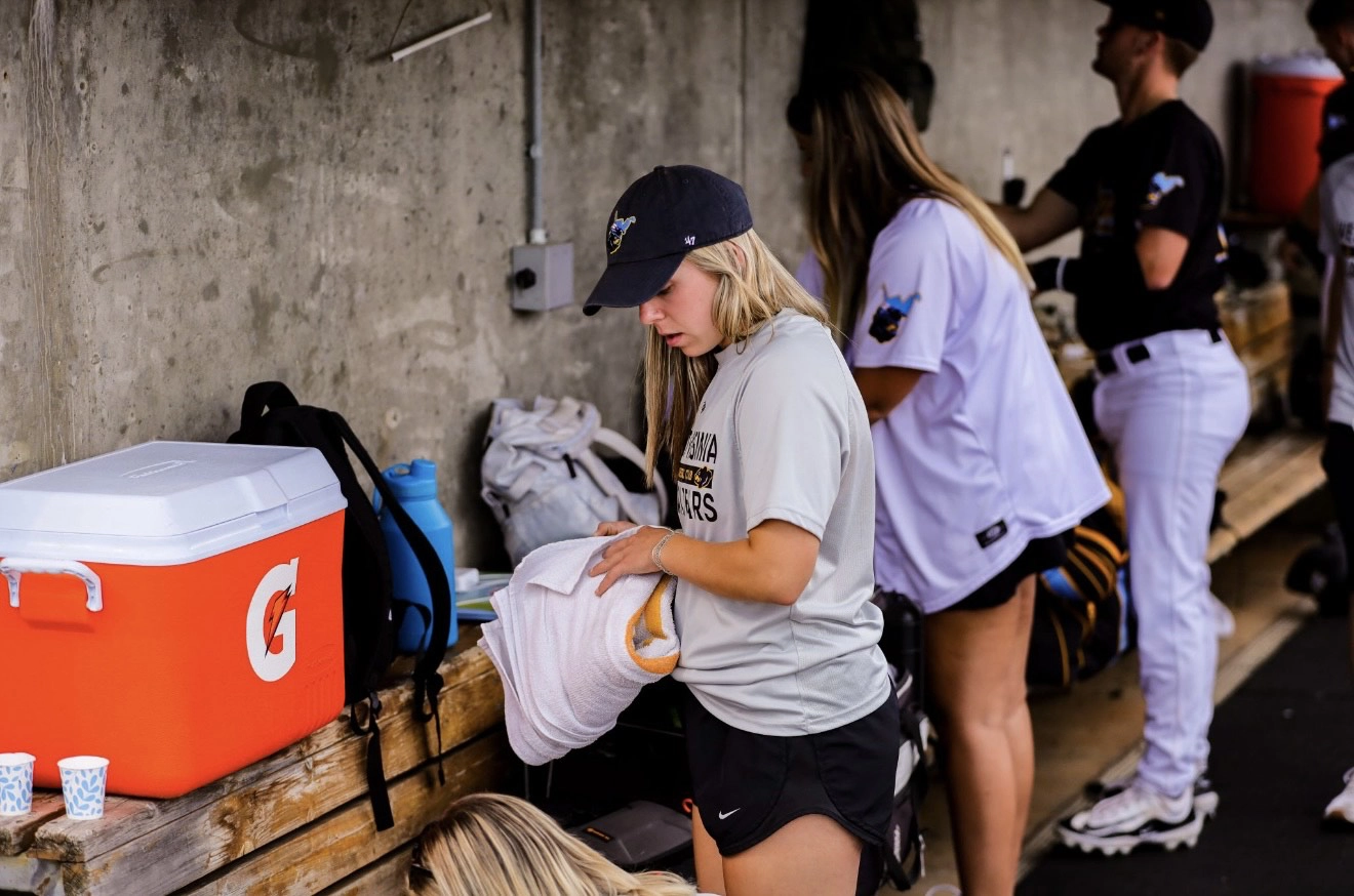 A person in a baseball dugout folds a towel next to a cooler while others organize equipment.