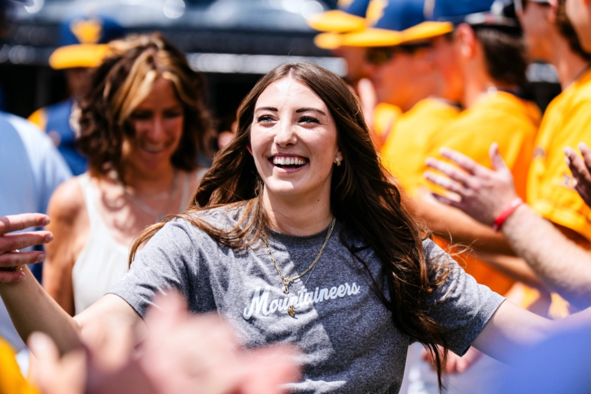 A smiling woman in a gray “Mountaineers” shirt walks forward with arms outstretched as people around her clap and greet her, with players in yellow uniforms in the background.