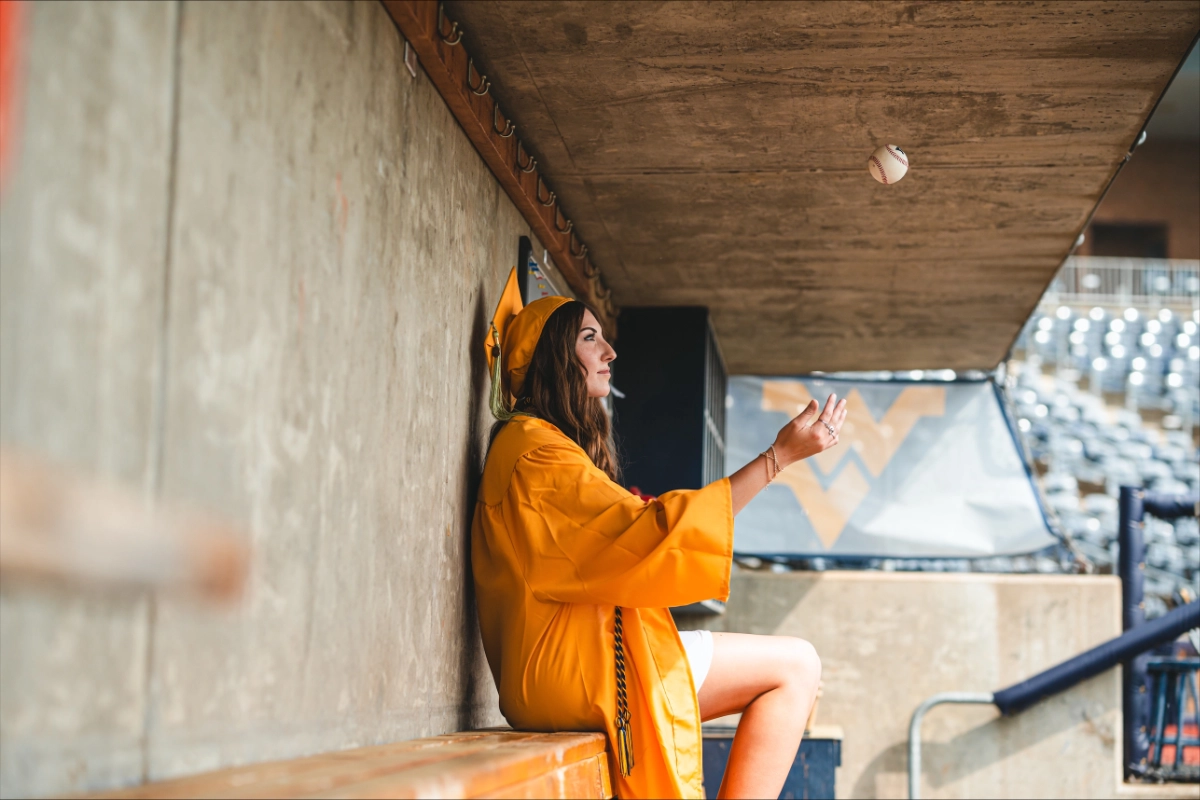 A graduate wearing a gold cap and gown sits on a dugout bench, casually tossing a baseball, with stadium seating and a WV logo visible in the background.