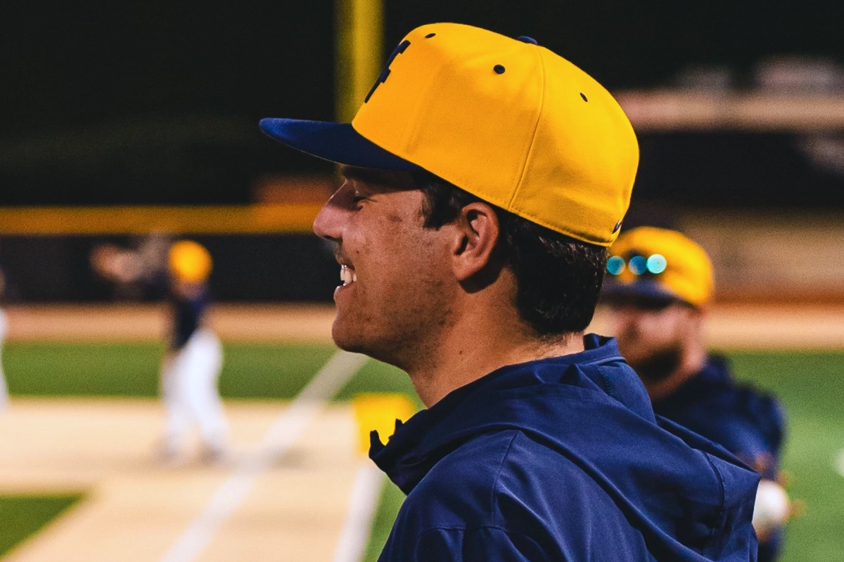 A baseball player in a yellow cap smiles while standing on the field.