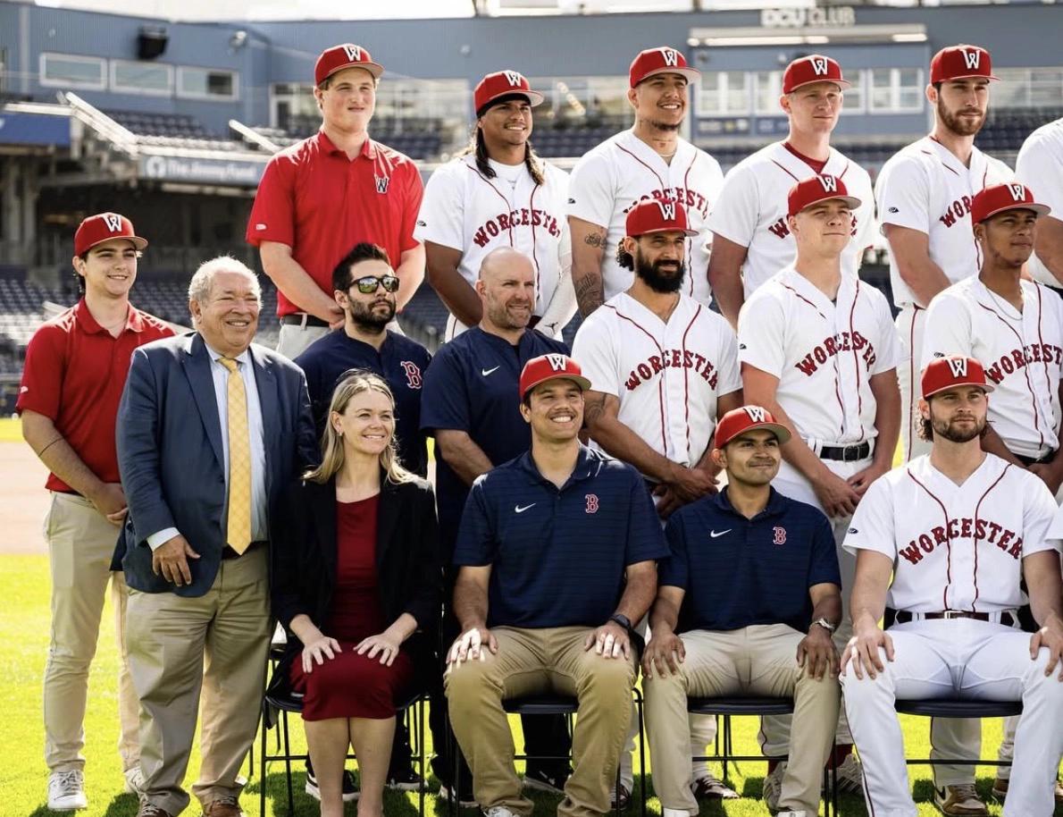 A baseball team in white uniforms and red caps poses for a group photo with coaches on a field.