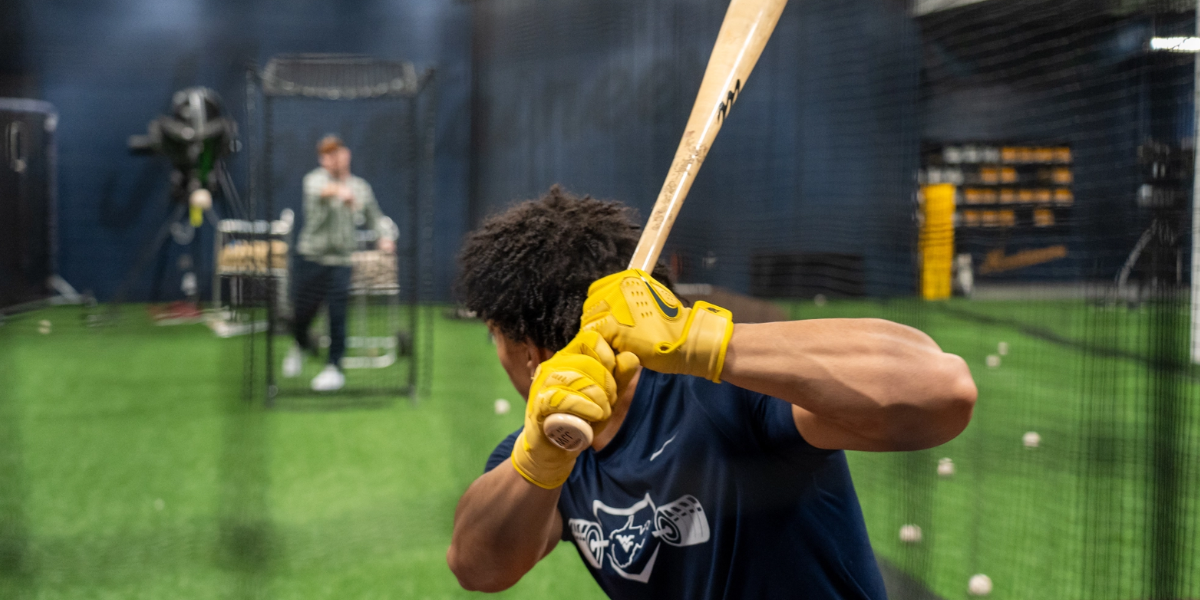 A baseball player wearing batting gloves prepares to swing in an indoor batting cage.