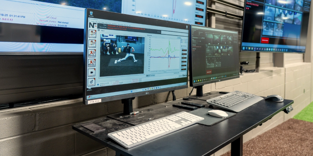 Multiple computer monitors display baseball motion analysis data and video in an indoor training facility.