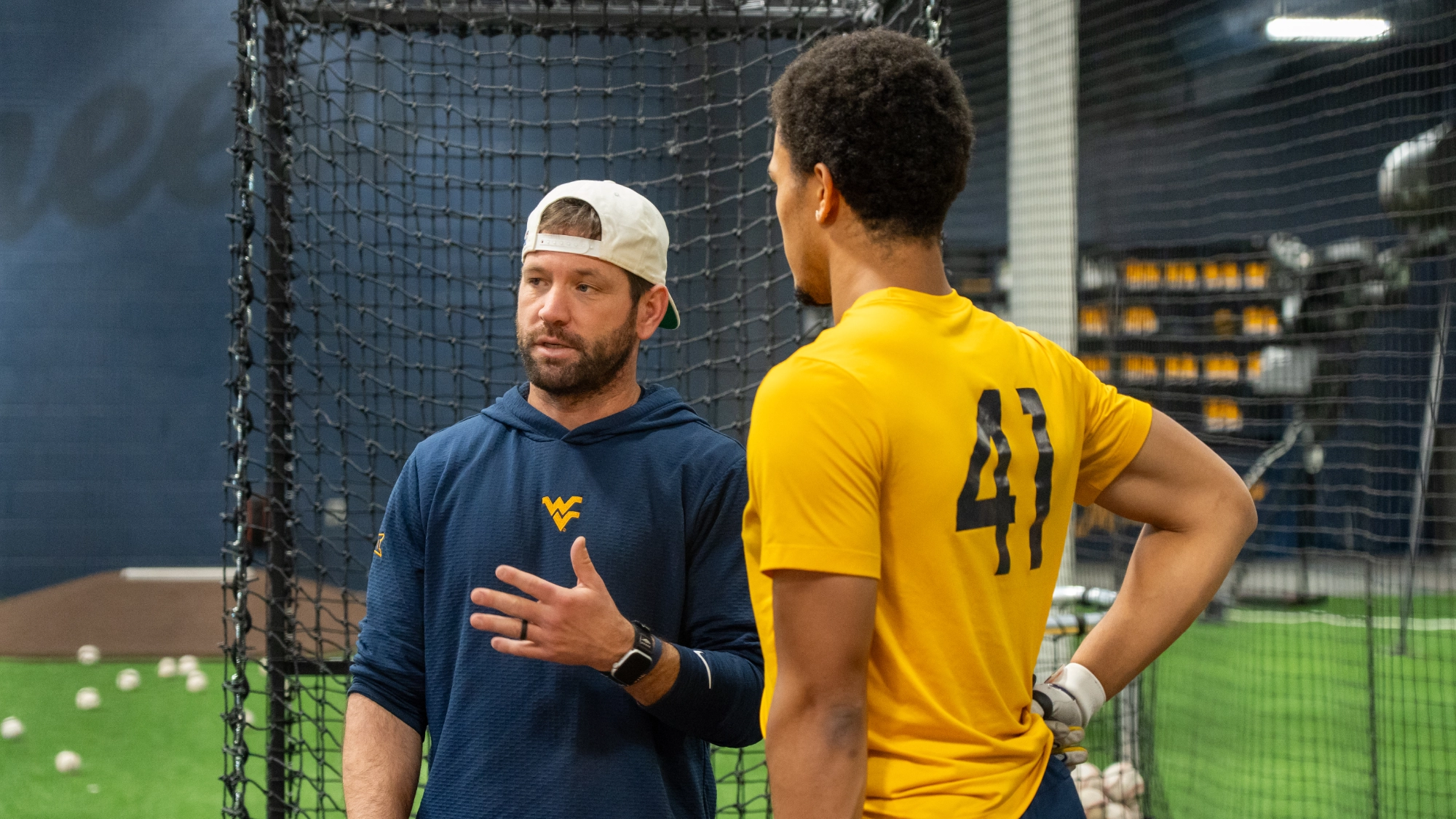A coach wearing a WVU hoodie speaks to a baseball player in a yellow shirt with the number 41, standing inside an indoor training facility with nets and baseballs visible.