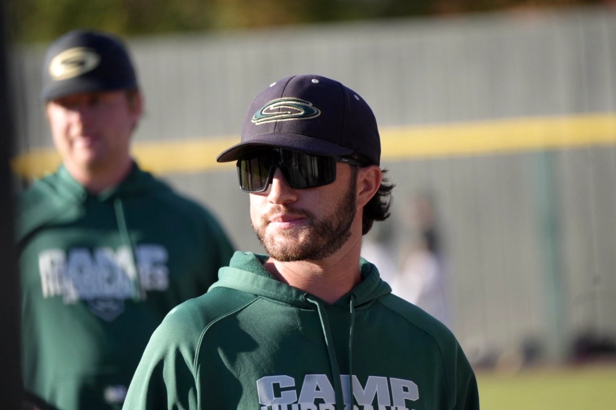 A baseball coach wearing sunglasses and a green hoodie stands on a field during practice.