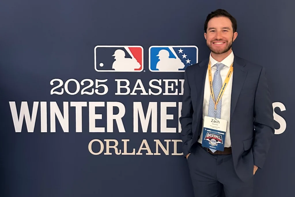 A man wearing a navy suit, tie, and event badge labeled &ldquo;Zach&rdquo; stands smiling in front of a wall displaying the MLB logo and &ldquo;2025 Baseball Winter Meetings Orlando.&rdquo;