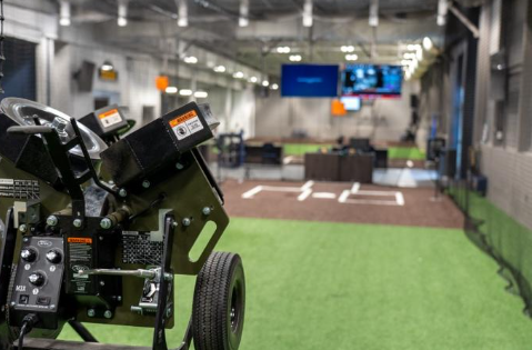A baseball pitching machine sits in the foreground of an indoor training facility with turf and batting lanes.